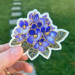May include: A sticker featuring a cluster of purple and blue flowers with gold accents. The flowers have a delicate, hand-painted style and are surrounded by green leaves.