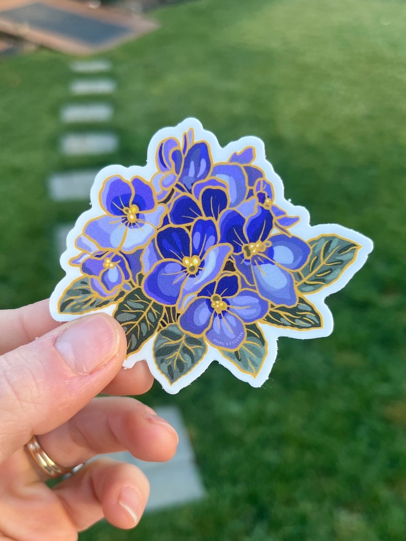 May include: A sticker featuring a cluster of purple and blue flowers with gold accents. The flowers have a delicate, hand-painted style and are surrounded by green leaves.