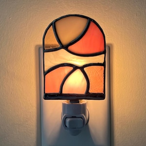 May include: A stained glass night light featuring an arched top and a geometric pattern of orange, peach, and cream-colored glass segments held together by dark metal solder lines. The light is plugged into a wall outlet and emits a warm glow.