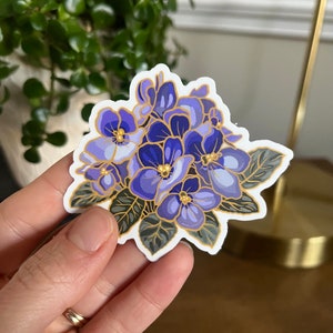 May include: A sticker featuring a cluster of purple and blue flowers with gold accents. The flowers are surrounded by green leaves.