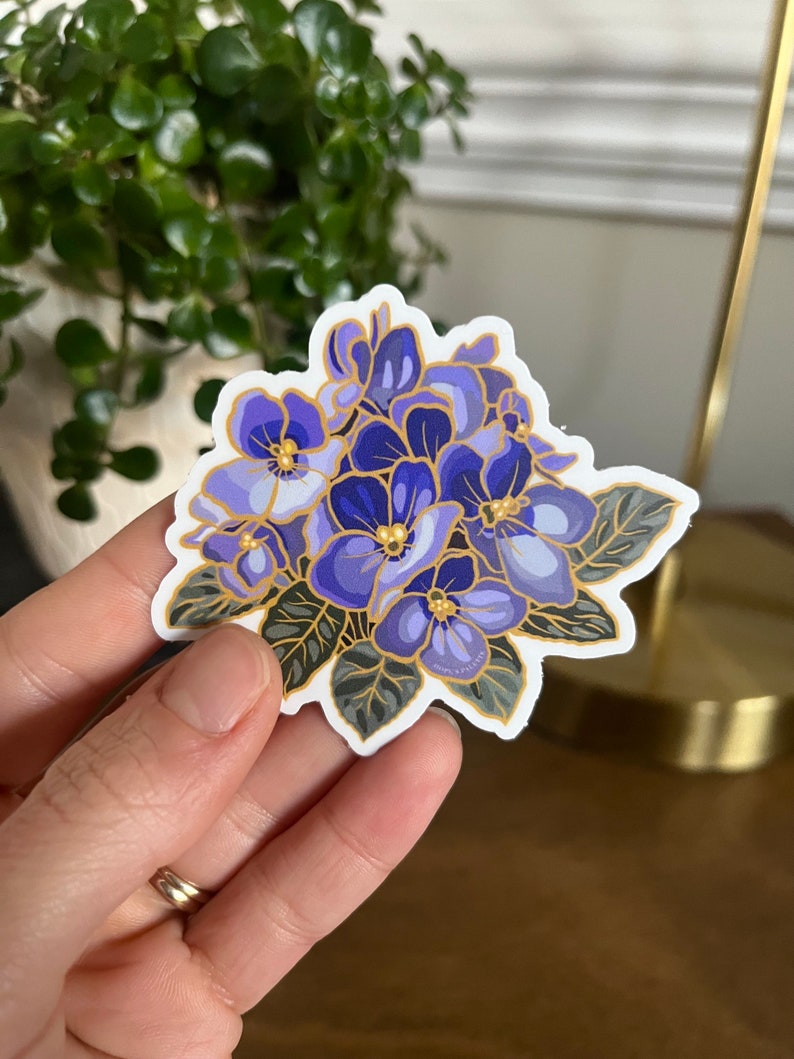 May include: A sticker featuring a cluster of purple and blue flowers with gold accents. The flowers are surrounded by green leaves.
