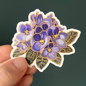 May include: A sticker featuring a cluster of purple and white flowers with gold accents. The flowers are surrounded by green leaves. The sticker has a white background and a white border.