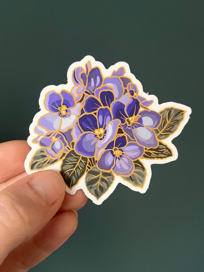 May include: A sticker featuring a cluster of purple and white flowers with gold accents. The flowers are surrounded by green leaves. The sticker has a white background and a white border.