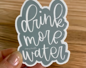 May include: Gray sticker with white lettering that reads "drink more water". The sticker has a white outline and a small white logo that reads "Hope's Palette".