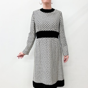 May include: A long-sleeved dress featuring a black and white checkerboard pattern. The dress has a black velvet band at the waist and a black velvet hem. The model is wearing black shoes and black leggings.
