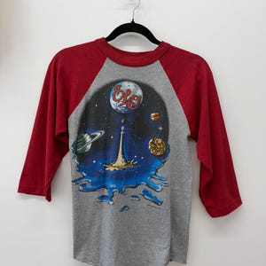 May include: A grey raglan t-shirt with three-quarter length red sleeves. The front features a graphic of a space scene with planets, a central spaceship, and the text ELO in red script. The design includes a blue liquid-like base at the bottom.