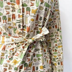 May include: A long-sleeved button-down shirt in a cream color featuring an all-over print of vintage paper doll clothing patterns in shades of green, orange, and yellow. The shirt includes a matching fabric tie belt at the waist.
