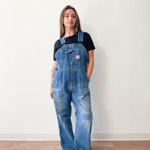 May include: Blue denim overalls with a chest pocket and a red and white logo patch. The overalls have a relaxed fit, with distressed details including a knee tear and a patch. The model is wearing a black t-shirt and white sneakers.