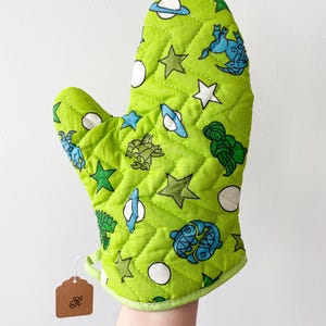 Op de afbeelding: Een limoengroene gewatteerde ovenwant met een patroon van witte sterren, planeten en blauwe en groene zeedieren. De want heeft een groene rand en een klein label.