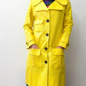 May include: A bright yellow, long coat with a textured, crocodile-like pattern. The coat features a collared neckline, large black buttons, and two front pockets. The coat is knee-length and has long sleeves.