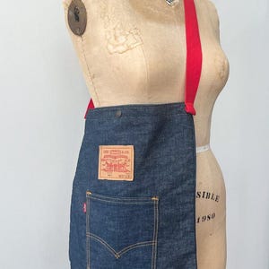 May include: A dark blue denim crossbody bag featuring a bright red strap and a rectangular tan leather patch with the text Levi Strauss & Co. Quality Clothing XX. The bag includes a front pocket with orange stitching and a small red tab label.