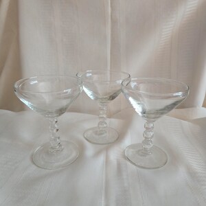 May include: Three clear champagne coupe glasses. Each glass has a round bowl, a stem with a series of small, round beads, and a circular base. The glasses are arranged on a white, textured surface.