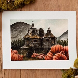 May include: A watercolor painting on paper depicting a stone cottage with two conical roofs nestled in a forest landscape with mountains in the background. Numerous orange pumpkins are scattered in the foreground near the cottage entrance.