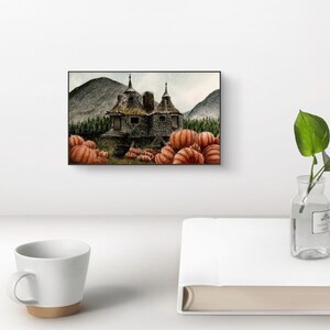 May include: A rectangular wall art print featuring a stone cottage with two pointed roofs, surrounded by a dense forest and mountains. The foreground is filled with numerous orange pumpkins. The artwork is displayed in a thin black frame.