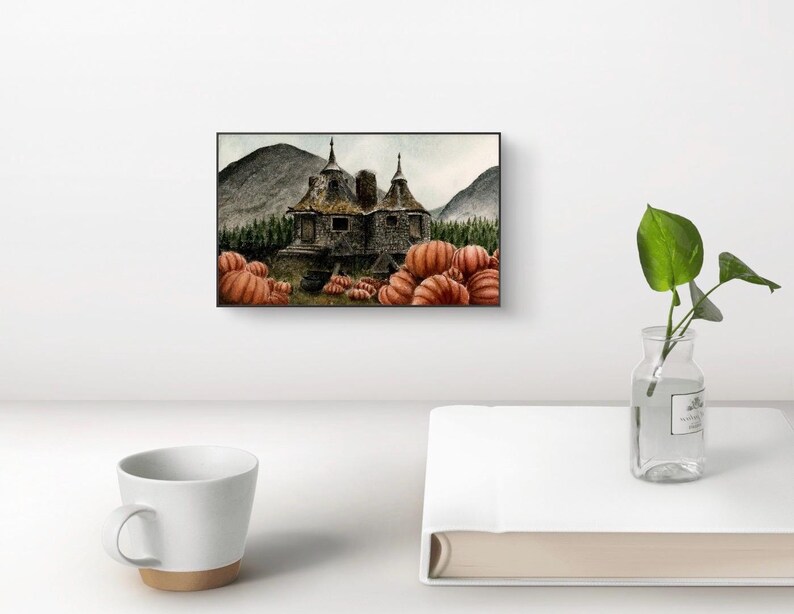 May include: A rectangular wall art print featuring a stone cottage with two pointed roofs, surrounded by a dense forest and mountains. The foreground is filled with numerous orange pumpkins. The artwork is displayed in a thin black frame.