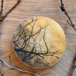May include: A round, hand-painted artwork featuring a spiderweb and tree branches. The artwork has a golden background with black branches and a white spiderweb. The artwork is set on a wooden surface with branches in the corners.