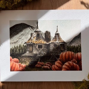 May include: A watercolor art print depicting a stone cottage with two conical roofs nestled in a forest landscape. The foreground features a large patch of orange pumpkins, and the background shows dark mountains under a muted, overcast sky.