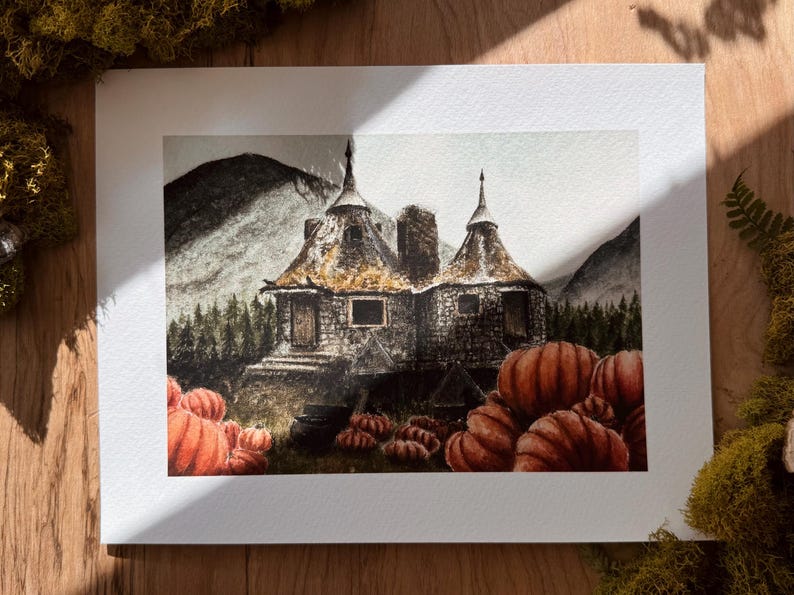 May include: A watercolor art print depicting a stone cottage with two conical roofs nestled in a forest landscape. The foreground features a large patch of orange pumpkins, and the background shows dark mountains under a muted, overcast sky.