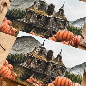 May include: Two identical rectangular art prints featuring a detailed illustration of a stone cottage with conical roofs, nestled among dark green pine trees and mountains, surrounded by a large patch of bright orange pumpkins in the foreground.