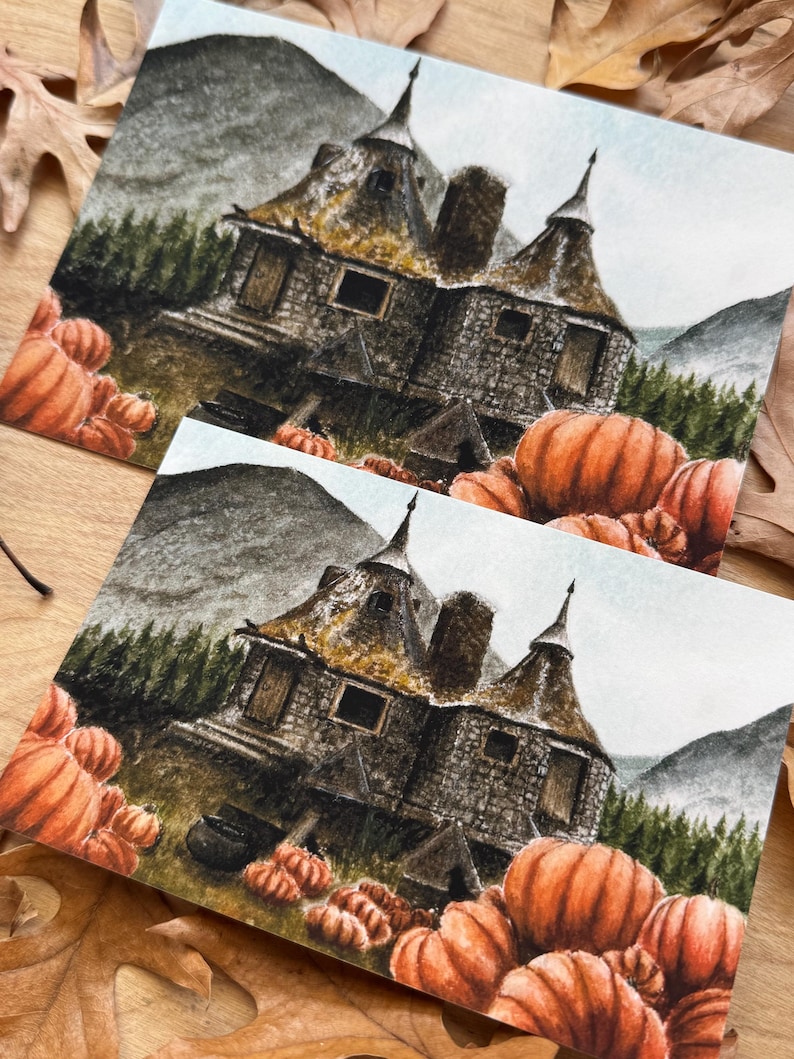 May include: Two identical rectangular art prints featuring a detailed illustration of a stone cottage with conical roofs, nestled among dark green pine trees and mountains, surrounded by a large patch of bright orange pumpkins in the foreground.