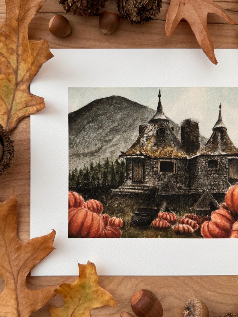 May include: A watercolor painting on white paper depicting a stone cottage with two conical roofs, situated in front of a dark mountain and pine forest. Numerous orange pumpkins are scattered in the foreground, along with a black cauldron.
