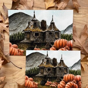 May include: Two rectangular art prints featuring a watercolor illustration of a stone cottage with two pointed roofs, surrounded by a dense forest, mountains, and a large patch of orange pumpkins in the foreground, set against a light sky.