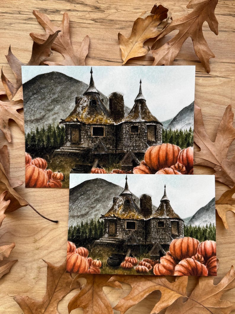 May include: Two rectangular art prints featuring a watercolor illustration of a stone cottage with two pointed roofs, surrounded by a dense forest, mountains, and a large patch of orange pumpkins in the foreground, set against a light sky.