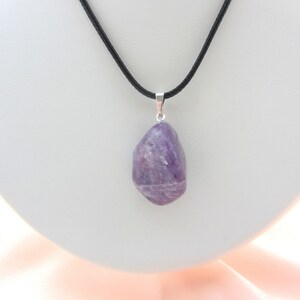 May include: A purple amethyst pendant hangs from a black cord necklace. The amethyst is a natural, irregularly shaped stone with a polished surface. The pendant is attached to a silver-colored bail. The necklace is displayed on a white surface.