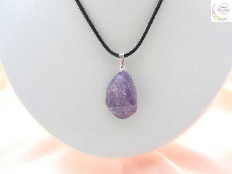 May include: A purple amethyst pendant hangs from a black cord necklace. The amethyst is a natural, irregularly shaped stone with a polished surface. The pendant is attached to a silver-colored bail. The necklace is displayed on a white surface.