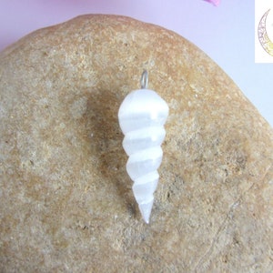 May include: White selenite crystal pendant in the shape of a spiral, hanging from a silver chain. The pendant is resting on a brown rock.