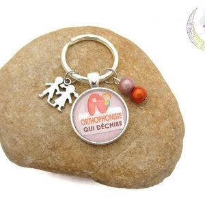 May include: Silver keyring with a pink and orange charm that says "Orthophoniste qui déchire". The charm is attached to a silver ring with two small silver figures of children.