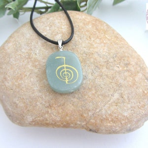 May include: A green stone pendant with a gold-coloured symbol on a black cord necklace. The pendant is shaped like a rounded rectangle and is resting on a light brown rock.