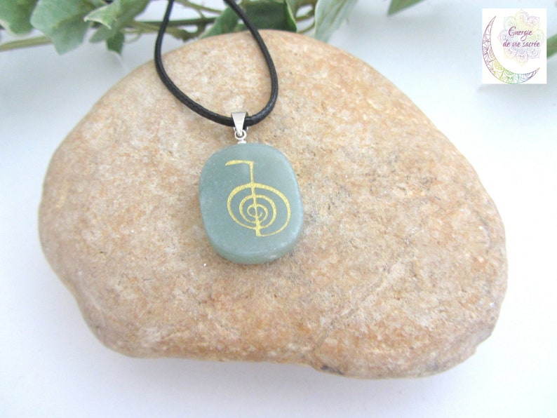 May include: A green stone pendant with a gold-coloured symbol on a black cord necklace. The pendant is shaped like a rounded rectangle and is resting on a light brown rock.