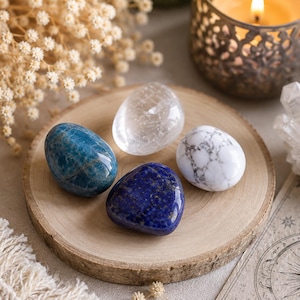May include: A collection of four smooth, polished stones resting on a circular wooden slice. The set includes one translucent clear quartz, one deep blue apatite, one dark blue lapis lazuli with gold flecks, and one white stone with grey veining.