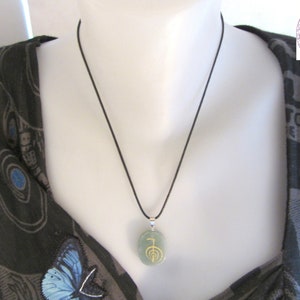May include: A green stone pendant necklace with a gold symbol on it. The necklace is on a black cord and is being worn by a person.