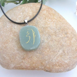 May include: A green stone pendant with a gold symbol on a black cord necklace. The pendant is oval shaped and the symbol is a stylized character.