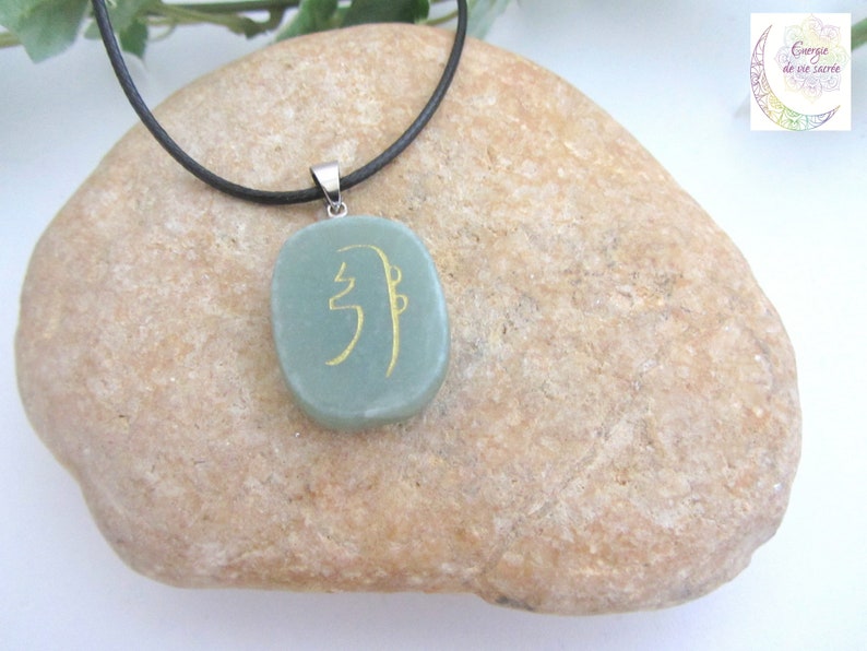 May include: A green stone pendant with a gold symbol on a black cord necklace. The pendant is oval shaped and the symbol is a stylized character.
