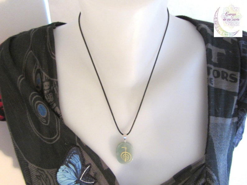 May include: A green stone pendant necklace with a gold symbol on it. The necklace is on a black cord and is being worn by a person.