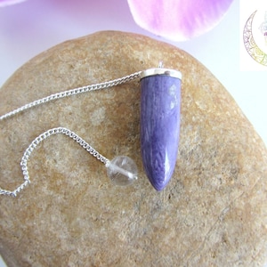 May include: A purple pendulum with a silver chain and a clear crystal sphere. The pendulum has a silver cap and a tapered, cylindrical shape. The chain is attached to the top of the pendulum and the crystal sphere. The pendulum is resting on a light brown stone.