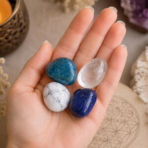 May include: Four polished, smooth, rounded stones resting in a palm. The collection includes one deep blue stone, one clear translucent stone, one white stone with grey veining, and one dark royal blue stone with subtle gold-colored flecks.