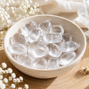May include: A collection of smooth, polished, transparent clear quartz stones contained within a round, speckled, off-white ceramic bowl. The stones are oval-shaped and exhibit natural internal fractures and varying degrees of clarity.