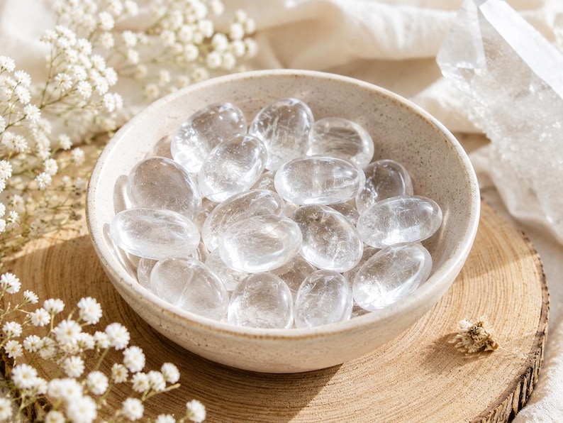 May include: A collection of smooth, polished, transparent clear quartz stones contained within a round, speckled, off-white ceramic bowl. The stones are oval-shaped and exhibit natural internal fractures and varying degrees of clarity.