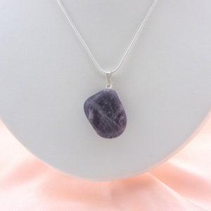 May include: A pendant necklace featuring a polished, irregularly shaped, purple amethyst stone. The stone is suspended from a delicate silver chain. The necklace is displayed on a white bust against a soft pink background.
