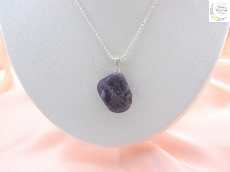 May include: A pendant necklace featuring a polished, irregularly shaped, purple amethyst stone. The stone is suspended from a delicate silver chain. The necklace is displayed on a white bust against a soft pink background.