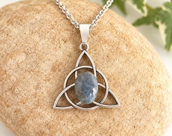 May include: A silver-toned metal pendant necklace featuring a triquetra knot design surrounding a smooth, oval-shaped blue-grey stone. The pendant hangs from a delicate silver-toned chain, showcasing a polished finish and intricate metalwork.