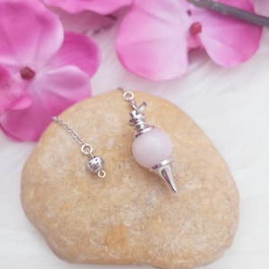 May include: A rose quartz pendulum with a silver-colored chain and accents rests on a beige stone. Pink orchid blossoms are in the background. The pendulum has a rounded, light pink stone at the top and a pointed tip.