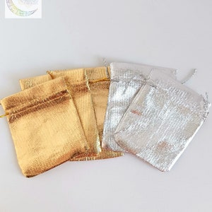 May include: A collection of small drawstring pouches featuring a metallic finish. Three pouches are gold and two are silver. Each pouch has a top closure with matching metallic drawstrings. A logo with a crescent moon and the text Energie de vie sacr&eacute;e.