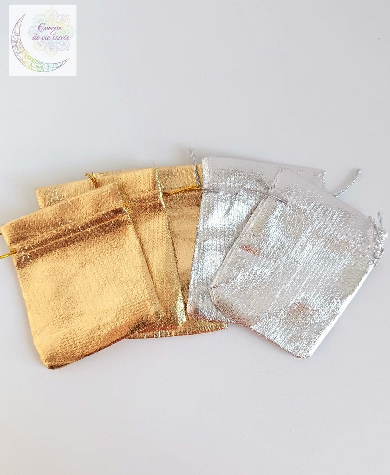 May include: A collection of small drawstring pouches featuring a metallic finish. Three pouches are gold and two are silver. Each pouch has a top closure with matching metallic drawstrings. A logo with a crescent moon and the text Energie de vie sacr&eacute;e.