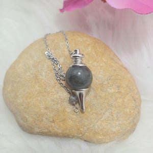 Op de afbeelding: Een zilverkleurige pendelketting met een donkergrijze, bolvormige steen. De pendel hangt aan een ketting en rust op een lichtbruine steen. Roze bloemen zijn op de achtergrond.