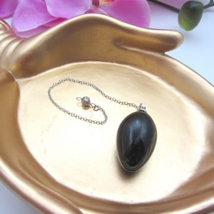 Könnte beinhalten: Schwarzes Pendel mit einer silbernen Kette und einer kleinen silbernen Kugel oben. Das Pendel liegt in einer goldfarbenen, handförmigen Schale.